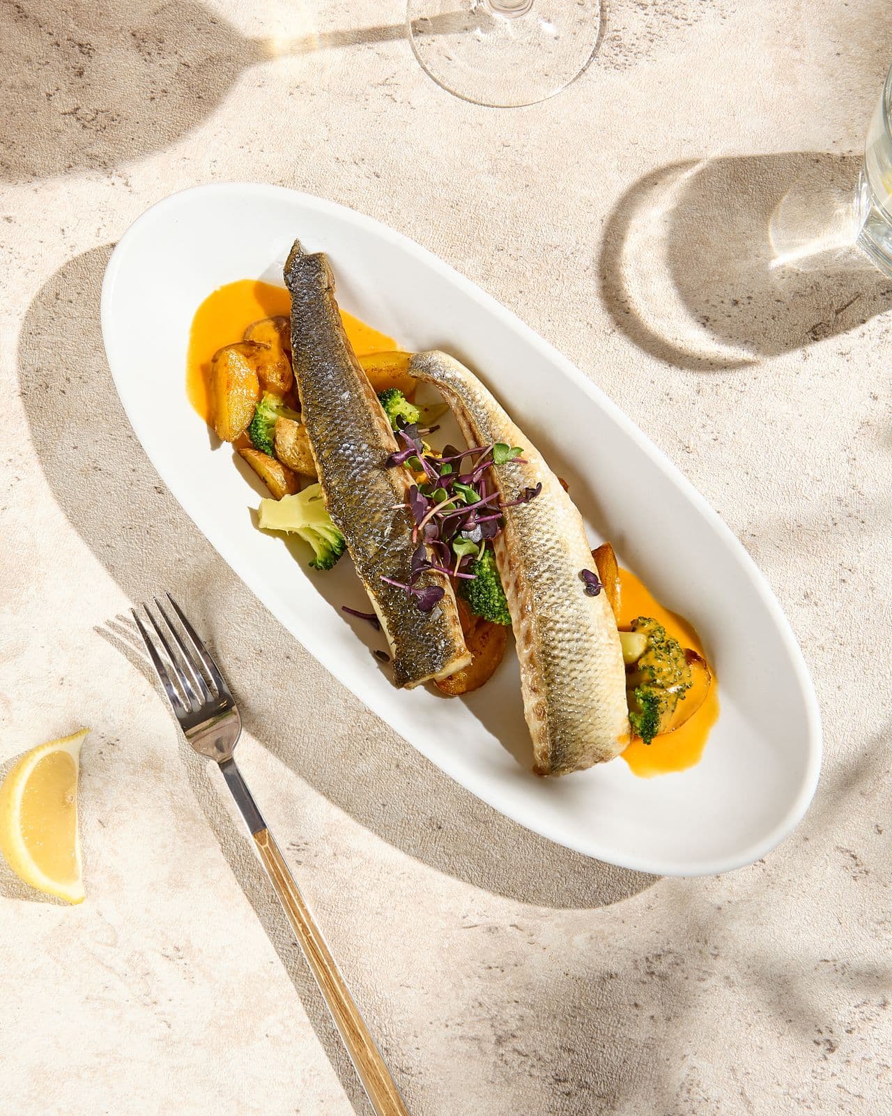 Two grilled fish fillets with potatoes, broccoli and an orange sauce on an oval plate, seen from above with a fork, a lemon wedge and a glass of white wine